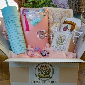 Puede incluir: Una caja de regalo de Rose of Lore Bookstore que contiene un vaso azul claro con pajita, un estuche rosa tipo sobre con borla, un diario floral, una vela con etiqueta de rosa y un llavero con un pequeño dije en forma de libro.