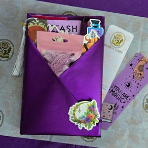 Può includere: Una confezione regalo viola con nappa, contenente vari oggetti come adesivi, un segnalibro con la scritta "YOU ARE MAGIC" e un sacchetto rosa. La scatola ha un adesivo floreale.