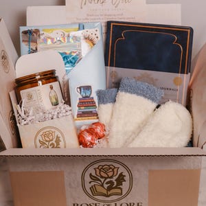 Peut inclure: Une boîte cadeau de la librairie Rose of Lore contenant un journal bleu foncé, une paire de chaussettes duveteuses blanches et bleues, une bougie en pot en verre avec étiquette, deux chocolats emballés en rouge et une carte de remerciement.
