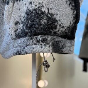 K&ouml;nnte beinhalten: Nahaufnahme einer grauen Fleecejacke mit schwarzem Spritzmuster. Die Jacke hat einen Kordelzug am unteren Saum. Der Stoff ist strukturiert und weich. Die Jacke wird auf einem wei&szlig;en St&auml;nder pr&auml;sentiert.