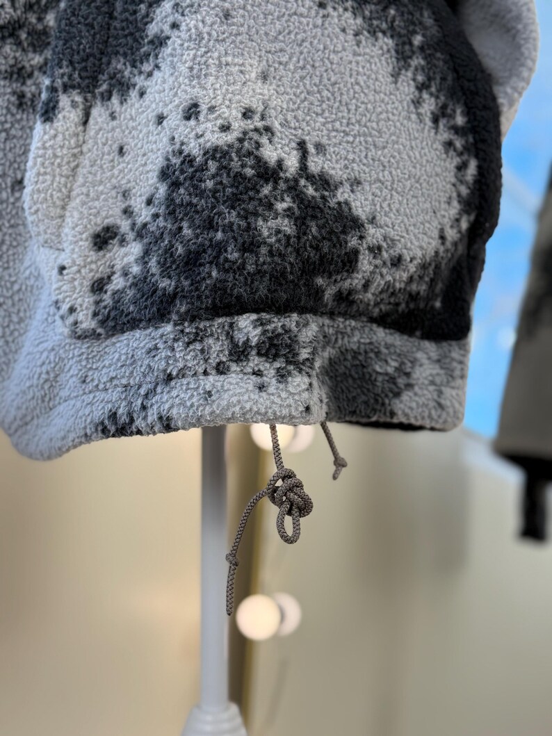 K&ouml;nnte beinhalten: Nahaufnahme einer grauen Fleecejacke mit schwarzem Spritzmuster. Die Jacke hat einen Kordelzug am unteren Saum. Der Stoff ist strukturiert und weich. Die Jacke wird auf einem wei&szlig;en St&auml;nder pr&auml;sentiert.