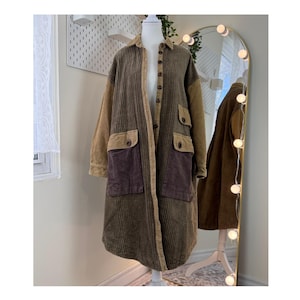 May include: A long, corduroy coat in shades of brown and tan, displayed on a mannequin. The coat features large pockets with button closures and contrasting panels of different colors. The coat has a collared neckline and button-down front.