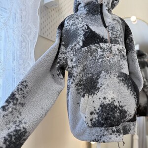 K&ouml;nnte beinhalten: Ein Kapuzenpullover aus grauem und schwarzem Fleece, der auf einer Schaufensterpuppe pr&auml;sentiert wird. Der Pullover hat einen halben Rei&szlig;verschluss, eine K&auml;ngurutasche und einen Kordelzug am Saum. Das Nike-Logo ist sichtbar.