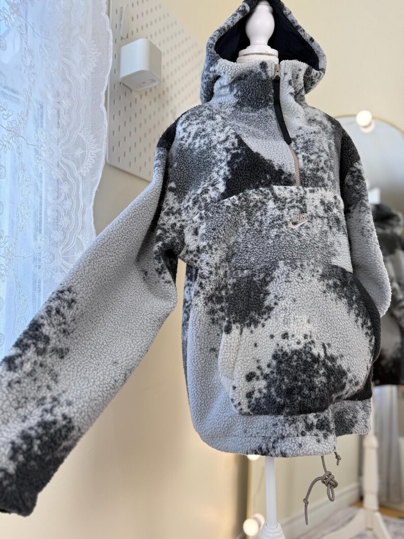 K&ouml;nnte beinhalten: Ein Kapuzenpullover aus grauem und schwarzem Fleece, der auf einer Schaufensterpuppe pr&auml;sentiert wird. Der Pullover hat einen halben Rei&szlig;verschluss, eine K&auml;ngurutasche und einen Kordelzug am Saum. Das Nike-Logo ist sichtbar.