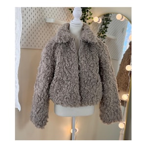 May include: A light brown faux fur jacket with a zip-up front and a wide collar. The jacket is displayed on a mannequin, showcasing its textured, curly fabric. The jacket is a cropped style, perfect for layering.