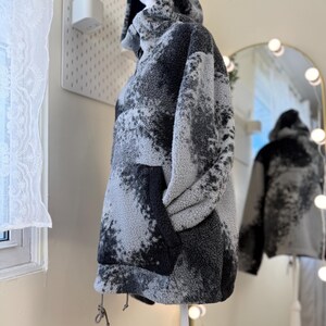 K&ouml;nnte beinhalten: Ein &uuml;bergro&szlig;er Fleece-Hoodie in Grau- und Schwarzt&ouml;nen, der auf einer Schaufensterpuppe pr&auml;sentiert wird. Die Jacke hat eine gro&szlig;e Vordertasche und einen Kordelzug am Saum. Das Muster &auml;hnelt einem Aquarelldesign.
