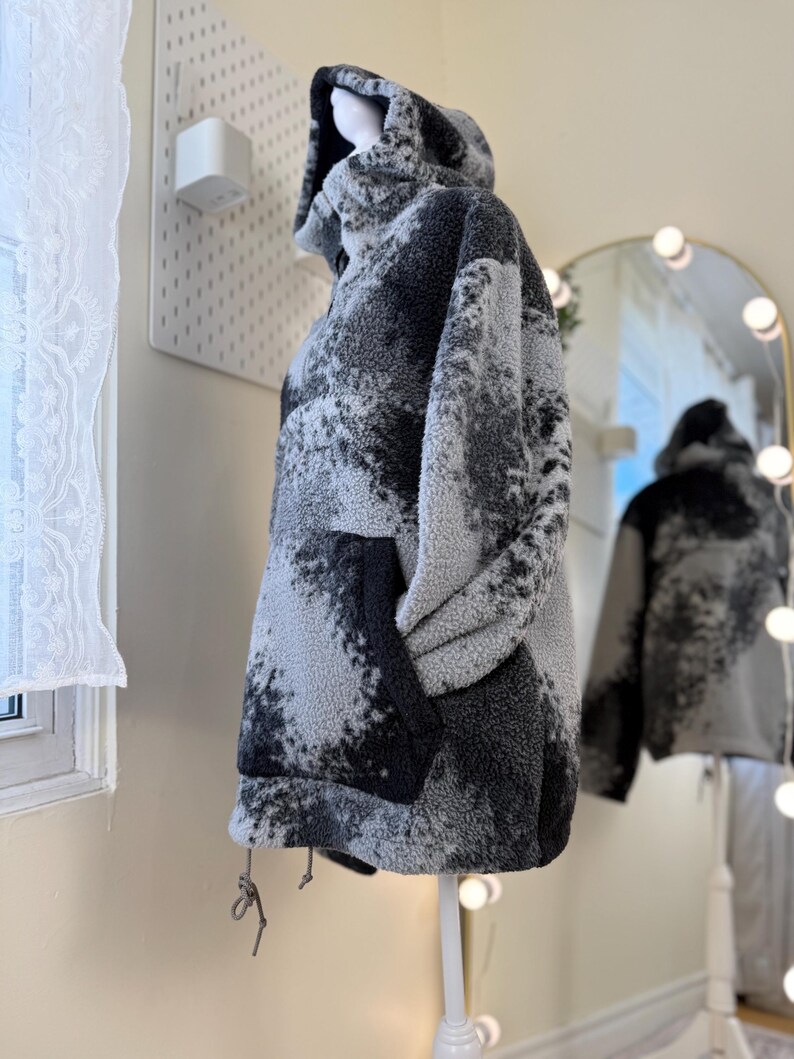 K&ouml;nnte beinhalten: Ein &uuml;bergro&szlig;er Fleece-Hoodie in Grau- und Schwarzt&ouml;nen, der auf einer Schaufensterpuppe pr&auml;sentiert wird. Die Jacke hat eine gro&szlig;e Vordertasche und einen Kordelzug am Saum. Das Muster &auml;hnelt einem Aquarelldesign.