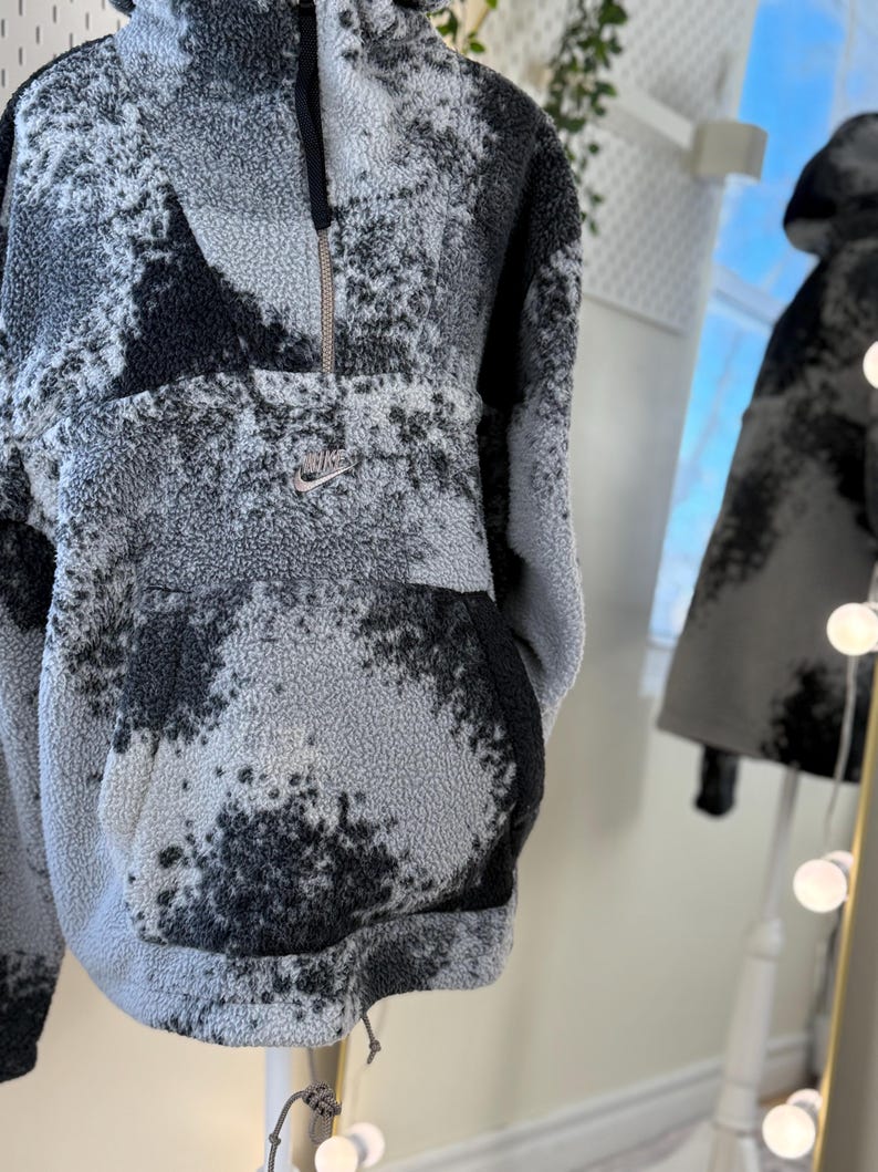 K&ouml;nnte beinhalten: Ein grau-schwarzer Fleece-Pullover mit einem Viertel-Rei&szlig;verschluss und einer Kapuze. Der Pullover hat ein einzigartiges Muster aus dunkelgrauen und schwarzen Flecken. Ein kleines Nike-Logo ist auf der Brust sichtbar.