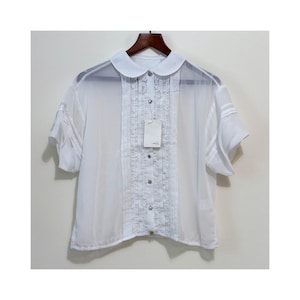 Puede incluir: Blusa blanca de manga corta con cuello Peter Pan y cierre de botones. La blusa tiene pliegues decorativos en la parte delantera y en las mangas. Una etiqueta está adjunta. La blusa está colgada en una percha de madera.