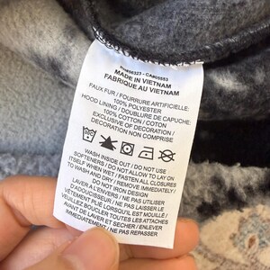 K&ouml;nnte beinhalten: Nahaufnahme eines Kleidungsst&uuml;cketiketts mit englischem und franz&ouml;sischem Text. Es zeigt "MADE IN VIETNAM" und listet Materialzusammensetzung und Pflegehinweise auf, einschlie&szlig;lich "WASH INSIDE OUT" und "DO NOT IRON DESIGN".