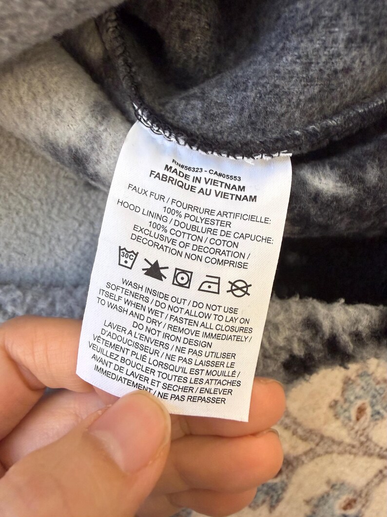 K&ouml;nnte beinhalten: Nahaufnahme eines Kleidungsst&uuml;cketiketts mit englischem und franz&ouml;sischem Text. Es zeigt "MADE IN VIETNAM" und listet Materialzusammensetzung und Pflegehinweise auf, einschlie&szlig;lich "WASH INSIDE OUT" und "DO NOT IRON DESIGN".