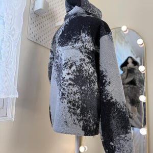 K&ouml;nnte beinhalten: Eine Kapuzenjacke aus grauem und schwarzem Fleece, pr&auml;sentiert auf einer Schaufensterpuppe. Die Jacke hat ein einzigartiges, gesprenkeltes Muster und ein weiches, strukturiertes Aussehen. Die R&uuml;ckansicht zeigt die Kapuze und die Gesamtform des Kleidungsst&uuml;cks.