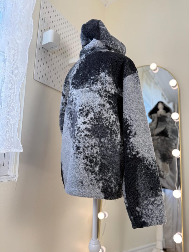 K&ouml;nnte beinhalten: Eine Kapuzenjacke aus grauem und schwarzem Fleece, pr&auml;sentiert auf einer Schaufensterpuppe. Die Jacke hat ein einzigartiges, gesprenkeltes Muster und ein weiches, strukturiertes Aussehen. Die R&uuml;ckansicht zeigt die Kapuze und die Gesamtform des Kleidungsst&uuml;cks.