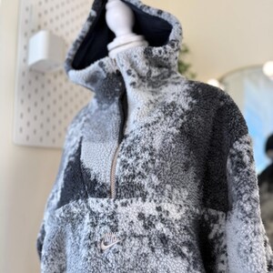 K&ouml;nnte beinhalten: Ein grauer und schwarzer Nike Fleece-Hoodie mit halbem Rei&szlig;verschluss und Kapuze. Der Hoodie hat ein kleines Nike-Logo auf der Brust. Das Fleecematerial hat ein gesprenkeltes Muster.