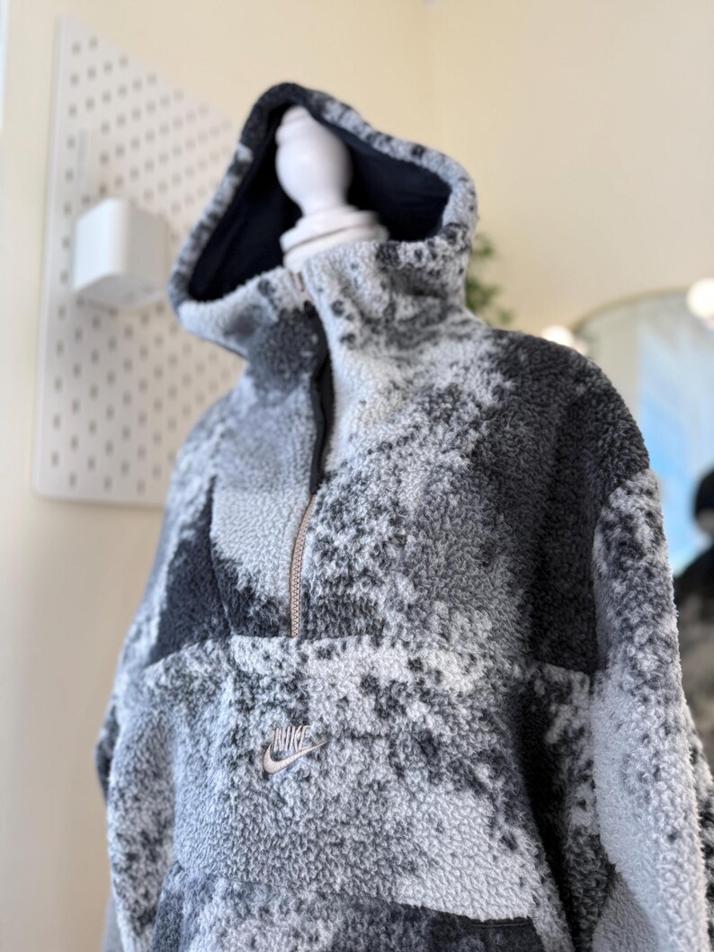 K&ouml;nnte beinhalten: Ein grauer und schwarzer Nike Fleece-Hoodie mit halbem Rei&szlig;verschluss und Kapuze. Der Hoodie hat ein kleines Nike-Logo auf der Brust. Das Fleecematerial hat ein gesprenkeltes Muster.