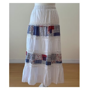 May include: A white, tiered skirt with a patchwork design. The skirt features alternating sections of white fabric and patterned patches in red, blue, and floral prints. Crochet lace trim separates the tiers. The skirt has an elasticated waistband.