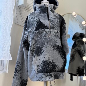 K&ouml;nnte beinhalten: Ein grau-schwarzer Fleece-Hoodie mit Kordelzug am Saum und einer Vordertasche. Der Hoodie hat einen hohen Kragen, eine Kapuze und einen halben Rei&szlig;verschluss. Das Fleece hat ein gesprenkeltes Muster und ein kleines Logo auf der Brust.