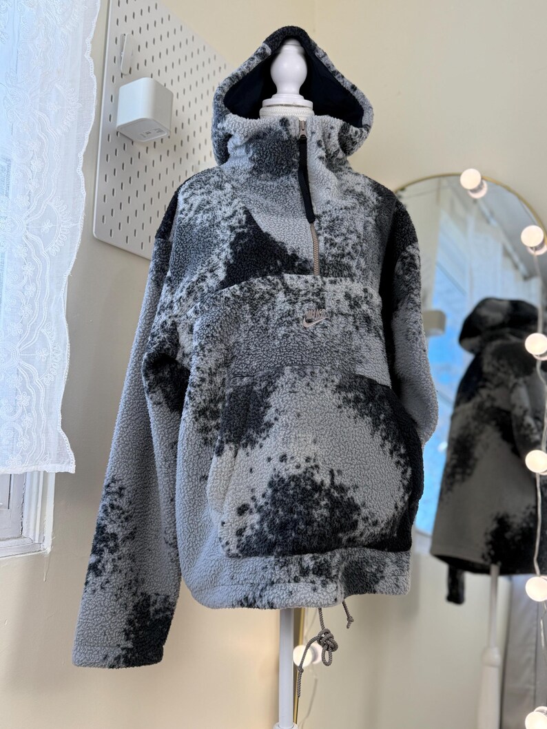 K&ouml;nnte beinhalten: Ein grau-schwarzer Fleece-Hoodie mit Kordelzug am Saum und einer Vordertasche. Der Hoodie hat einen hohen Kragen, eine Kapuze und einen halben Rei&szlig;verschluss. Das Fleece hat ein gesprenkeltes Muster und ein kleines Logo auf der Brust.