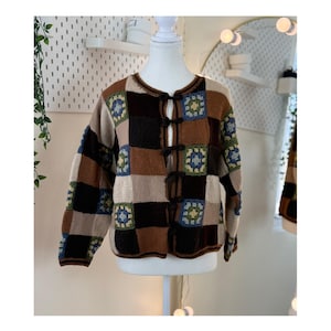 Peut inclure: Cardigan patchwork aux tons marron, beige, noir et bleu, présenté sur un mannequin. Le pull a une encolure ronde et se ferme avec quatre brandebourgs marron foncé. Le design intègre des motifs carrés au crochet.