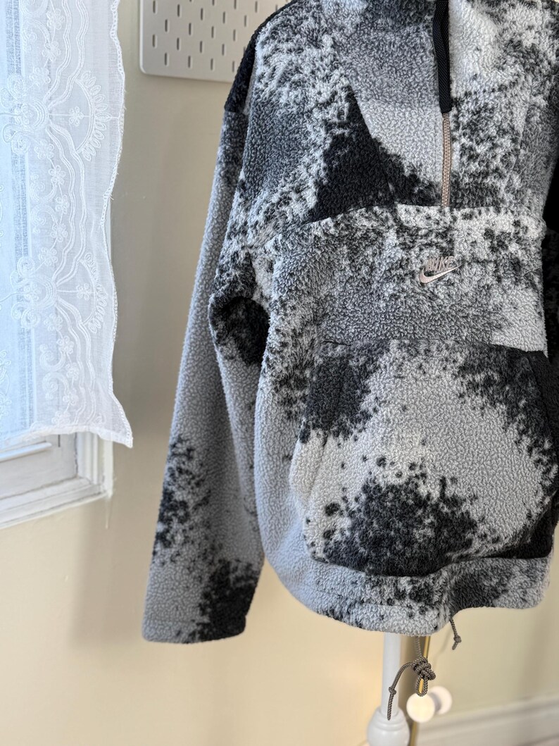 K&ouml;nnte beinhalten: Grau-schwarzer Fleece-Pullover mit Viertelrei&szlig;verschluss und K&auml;ngurutasche. Der Pullover hat ein gesprenkeltes Muster und ein kleines Nike-Logo auf der Brust. Der Kordelzug am Saum erm&ouml;glicht eine individuelle Passform.