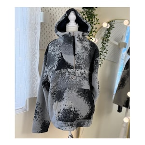 K&ouml;nnte beinhalten: Ein grauer und schwarzer Fleece-Hoodie mit einem halben Rei&szlig;verschluss und einer Kapuze. Der Hoodie hat ein einzigartiges Muster aus dunkelgrauen und schwarzen Flecken auf einem hellgrauen Hintergrund. Das Kleidungsst&uuml;ck wird auf einer Schaufensterpuppe pr&auml;sentiert.