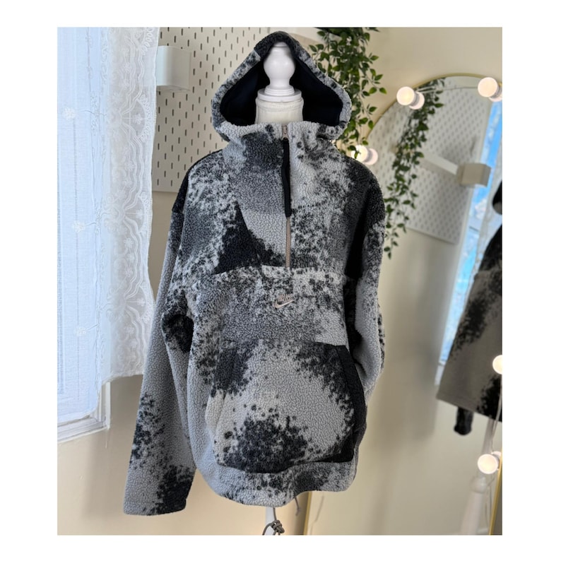 K&ouml;nnte beinhalten: Ein grauer und schwarzer Fleece-Hoodie mit einem halben Rei&szlig;verschluss und einer Kapuze. Der Hoodie hat ein einzigartiges Muster aus dunkelgrauen und schwarzen Flecken auf einem hellgrauen Hintergrund. Das Kleidungsst&uuml;ck wird auf einer Schaufensterpuppe pr&auml;sentiert.