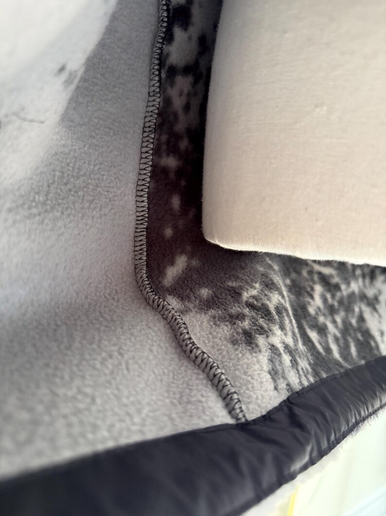 K&ouml;nnte beinhalten: Nahaufnahme eines grau-wei&szlig; gemusterten Fleece-Stoffs mit schwarzem Besatz. Der Stoff hat eine weiche, pl&uuml;schige Textur und ist mit schwarzem Faden zusammengen&auml;ht. Ein wei&szlig;es, zylindrisches Objekt ist teilweise sichtbar.