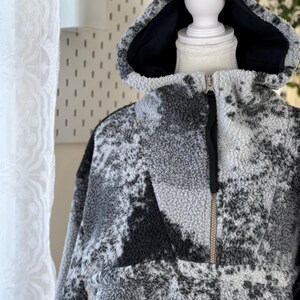 K&ouml;nnte beinhalten: Ein grau-schwarzer Fleece-Hoodie mit einem halben Rei&szlig;verschluss und einem schwarzen Innenfutter in der Kapuze. Das Nike-Logo ist unten auf der Vorderseite sichtbar. Der Hoodie hat ein strukturiertes Muster und wird auf einer Schaufenquin pr&auml;sentiert.