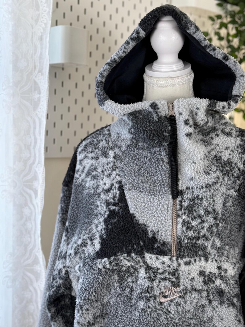 K&ouml;nnte beinhalten: Ein grau-schwarzer Fleece-Hoodie mit einem halben Rei&szlig;verschluss und einem schwarzen Innenfutter in der Kapuze. Das Nike-Logo ist unten auf der Vorderseite sichtbar. Der Hoodie hat ein strukturiertes Muster und wird auf einer Schaufenquin pr&auml;sentiert.