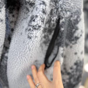 K&ouml;nnte beinhalten: Nahaufnahme einer grau-wei&szlig;en Fleecejacke mit einer schwarzen Rei&szlig;verschlusstasche. Die Jacke hat ein gesprenkeltes Muster und ein weiches, strukturiertes Aussehen. Das Fleecematerial ist dick und warm, geeignet f&uuml;r kaltes Wetter.