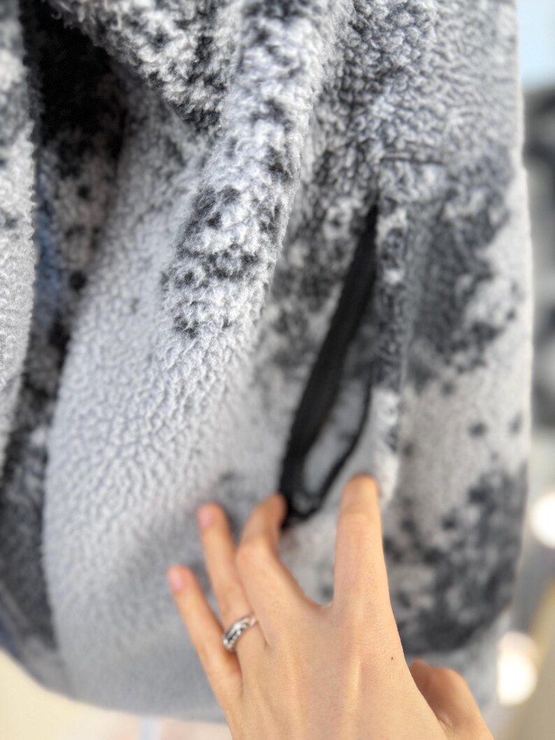 K&ouml;nnte beinhalten: Nahaufnahme einer grau-wei&szlig;en Fleecejacke mit einer schwarzen Rei&szlig;verschlusstasche. Die Jacke hat ein gesprenkeltes Muster und ein weiches, strukturiertes Aussehen. Das Fleecematerial ist dick und warm, geeignet f&uuml;r kaltes Wetter.