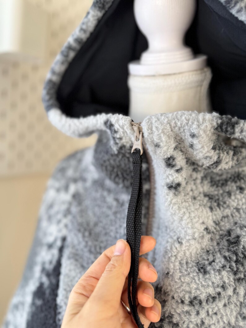 K&ouml;nnte beinhalten: Nahaufnahme einer Kapuzenjacke mit einem schwarz-wei&szlig;en Muster. Die Jacke hat ein schwarzes Innenfutter und einen Rei&szlig;verschluss mit einem schwarzen Zugband. Der Stoff scheint ein weiches, strukturiertes Material zu sein.