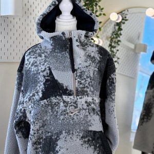 K&ouml;nnte beinhalten: Ein grau-schwarzer Fleece-Hoodie mit Kapuze und halbem Rei&szlig;verschluss. Der Hoodie hat ein gesprenkeltes Muster und ein kleines Nike-Logo auf der Brust. Die Kapuze hat ein schwarzes Futter.