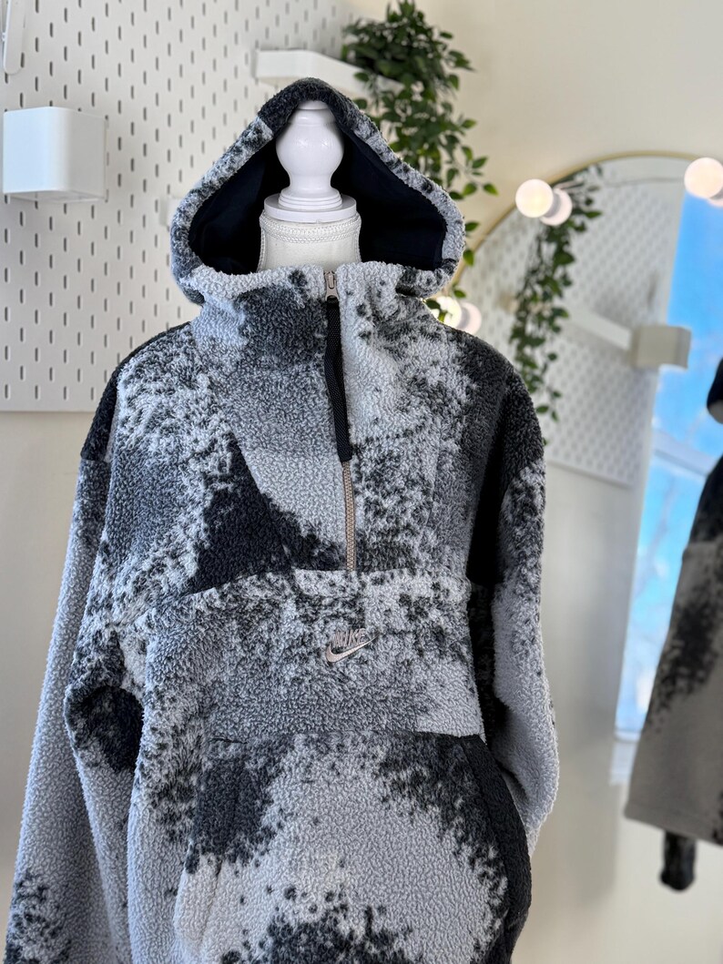 K&ouml;nnte beinhalten: Ein grau-schwarzer Fleece-Hoodie mit Kapuze und halbem Rei&szlig;verschluss. Der Hoodie hat ein gesprenkeltes Muster und ein kleines Nike-Logo auf der Brust. Die Kapuze hat ein schwarzes Futter.