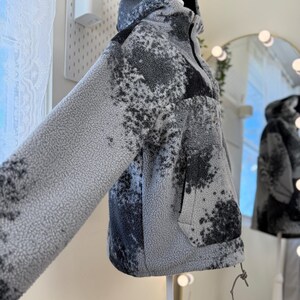 K&ouml;nnte beinhalten: Eine Kapuzenjacke mit einem abstrakten Muster in Grau und Schwarz. Die Jacke hat einen Kordelzug am Saum und eine Vordertasche. Die Jacke wird auf einer Schaufensterpuppe pr&auml;sentiert. Die Jacke ist aus einem weichen, strukturierten Material gefertigt.