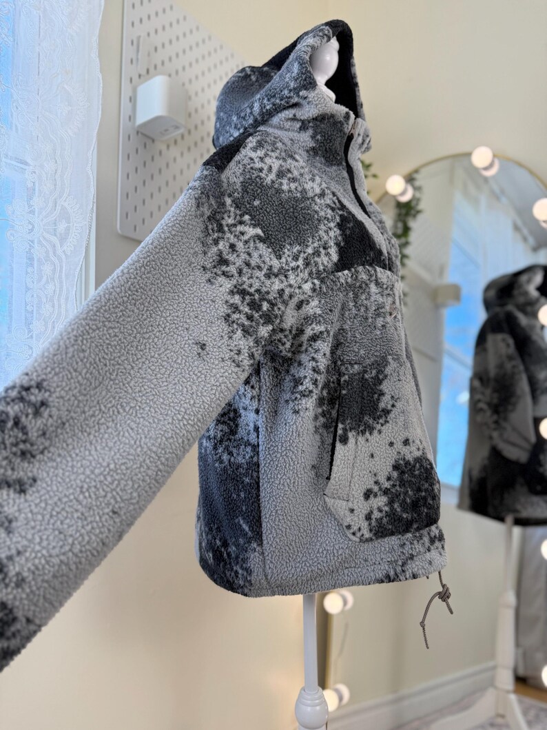 K&ouml;nnte beinhalten: Eine Kapuzenjacke mit einem abstrakten Muster in Grau und Schwarz. Die Jacke hat einen Kordelzug am Saum und eine Vordertasche. Die Jacke wird auf einer Schaufensterpuppe pr&auml;sentiert. Die Jacke ist aus einem weichen, strukturierten Material gefertigt.