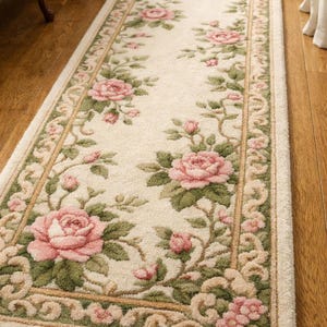 Puede incluir: Alfombra de pasillo color crema con un estampado floral de rosas. La alfombra presenta rosas rosas, hojas verdes y un borde decorativo en tonos beige y verde. La alfombra es rectangular y está colocada sobre un suelo de madera.