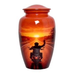 May include: A cylindrical metal cremation urn with a glossy finish featuring a printed graphic of a motorcyclist riding down a road towards a bright sunset. The urn has a rounded lid and a tapered base, displaying warm orange and red tones.