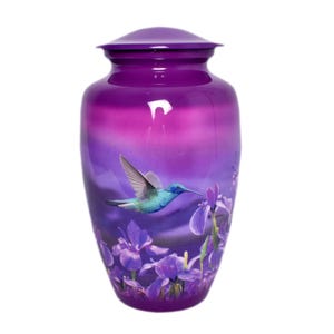 May include: A cylindrical metal cremation urn featuring a vibrant purple gradient finish. The exterior is decorated with a detailed graphic of a teal hummingbird in flight hovering near a cluster of purple iris flowers against a soft purple background.