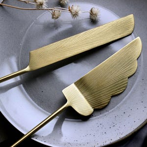 May include: Two gold-coloured serving utensils with textured surfaces rest on a grey plate. One utensil is a cake server with a scalloped edge, and the other is a rectangular knife. The utensils have long, thin handles.