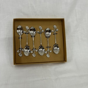 Könnte beinhalten: Ein Set aus sechs silberfarbenen Löffeln mit floralen Designs an den Griffen, präsentiert in einer braunen rechteckigen Box. Die Löffel haben eine klassische tropfenförmige Schale. Die floralen Details verleihen dem Besteck eine dekorative Note.