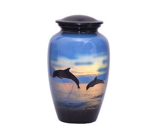 May include: A cylindrical cremation urn with a glossy black lid and base. The body features a printed scene of two dolphins leaping from the ocean against a sunset sky with gradients of blue, orange, and yellow hues across the surface.