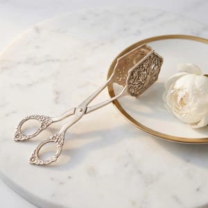Puede incluir: Pinzas de servicio de metal plateado con grabados florales ornamentales en los mangos y en los extremos de agarre. Las pinzas tienen dos anillos circulares para los dedos y un mecanismo de bisagra, situadas junto a una flor blanca.
