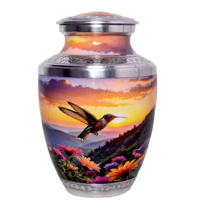 Könnte beinhalten: Dekorative Urne mit einer Sonnenuntergangsszene und einem fliegenden Kolibri. Die Urne hat einen silbernen Deckel und Boden, mit einem farbenfrohen Bild eines Kolibris, Bergen und Blumen. Der Hintergrund zeigt einen lebendigen Sonnenuntergang.