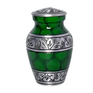 May include: A small, green urn with a silver-colored lid and decorative bands. The urn has a textured, mottled green surface and silver bands with floral and geometric patterns. The urn is designed to hold a small amount of cremated remains.