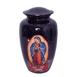 May include: A dark blue cremation urn with a speckled finish. The urn features an image of Our Lady of Guadalupe, with a halo and floral accents. A religious memorial item.