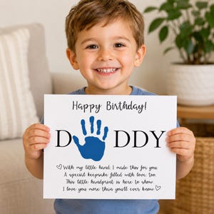 Könnte beinhalten: Ein weißes rechteckiges Schild mit schwarzem Text Happy Birthday! DADDY, bei dem ein blauer Handabdruck den Buchstaben A ersetzt. Darunter steht in schwarzer Schrift With my little hand, I made this for you. A special keepsake filled with love, too.