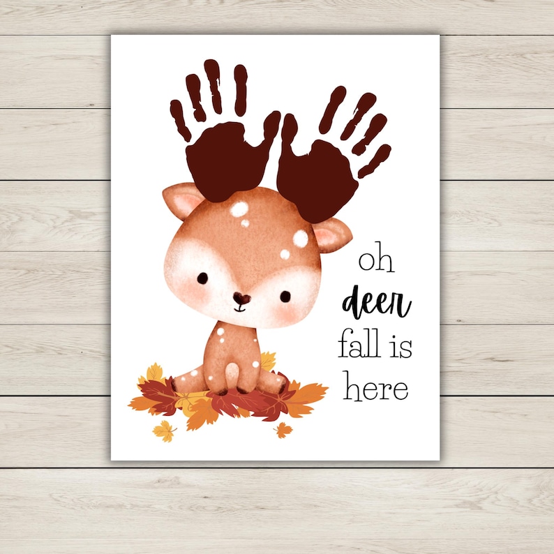 Puede incluir: Una impresi&oacute;n caprichosa con un ciervo de dibujos animados con astas de huellas de manos, sentado sobre hojas de oto&ntilde;o. El texto "oh deer fall is here" est&aacute; en negro. Fondo blanco.