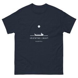 May include: Navy blue t-shirt featuring a white graphic of a ship, a moon, and the words "skeleton coast" and "Namibia". The design is minimalist and the shirt is made of a soft material.