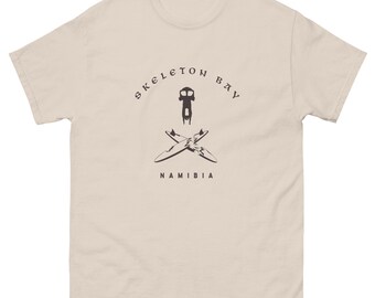 May include: A light beige t-shirt with a graphic design. The design features the words "Skeleton Bay" arched above a central image of a stylized figure and crossed surfboards, with "Namibia" below. The shirt has short sleeves and a crew neck.